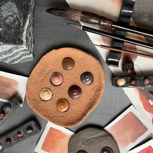 Puede incluir: Una paleta de terracota con seis pocillos de pintura llenos de diferentes tonos de marrón, rojo y naranja. La paleta está rodeada de varias muestras de pintura y pinceles.