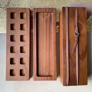 May include: Three wooden boxes of varying designs. The first has square cutouts, the second is a tray, and the third is wrapped with leather straps and a metal ring. All are made of a warm brown wood.