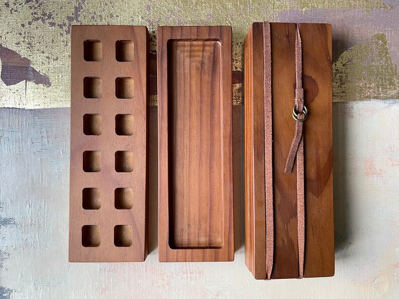 May include: Three wooden boxes of varying designs. The first has square cutouts, the second is a tray, and the third is wrapped with leather straps and a metal ring. All are made of a warm brown wood.