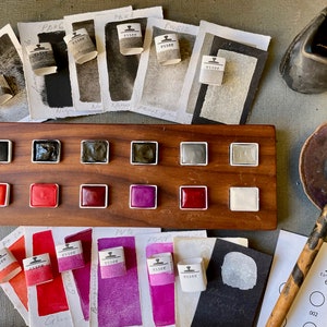 May include: A wooden palette with watercolor paints in various colors, including black, red, and purple. The palette is surrounded by watercolor paper swatches and small paper rolls labeled "essee". A ceramic inkwell and a calligraphy pen are also present.