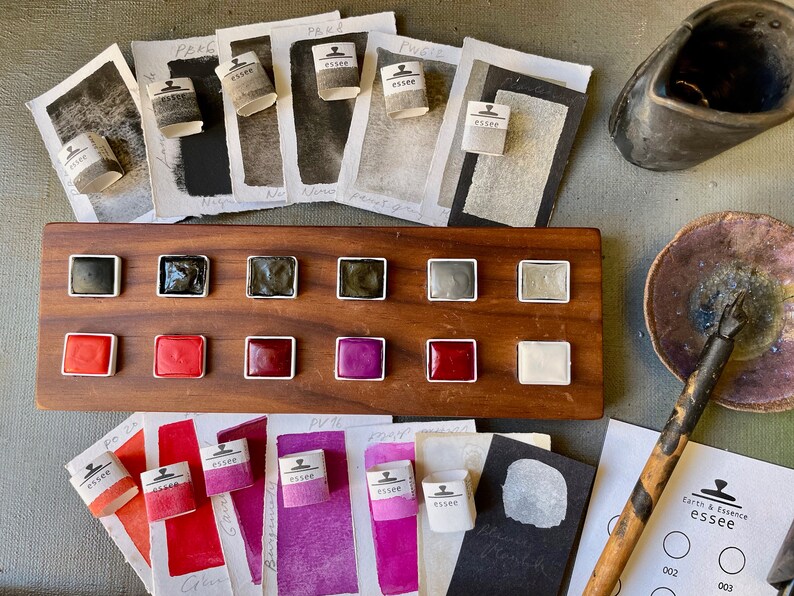 May include: A wooden palette with watercolor paints in various colors, including black, red, and purple. The palette is surrounded by watercolor paper swatches and small paper rolls labeled "essee". A ceramic inkwell and a calligraphy pen are also present.