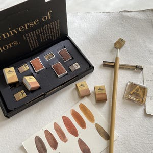 May include: A black box set with the text "A Universe of colors." contains watercolour paints in various shades of brown and beige. A paintbrush, colour swatches, and a small card are also present.