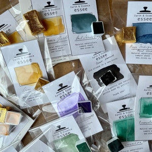 May include: An assortment of watercolour paint pans in various colours, including gold, green, purple, blue, and black. Each pan is labelled with the brand name "Earth & Essence essee" and a description of the colour. The pans are packaged in clear plastic bags with white labels.