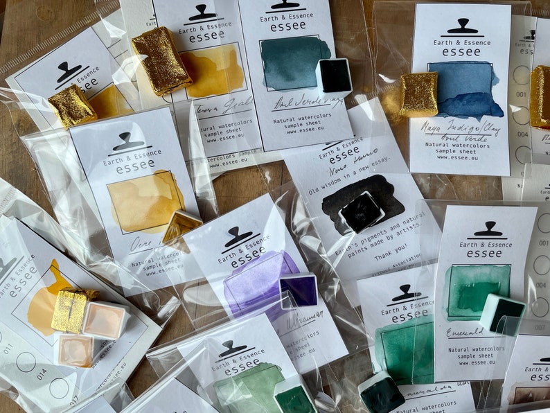 May include: An assortment of watercolour paint pans in various colours, including gold, green, purple, blue, and black. Each pan is labelled with the brand name "Earth & Essence essee" and a description of the colour. The pans are packaged in clear plastic bags with white labels.