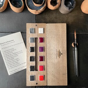 May include: A wooden watercolor palette with 12 color squares, a paintbrush, and a card listing the colors. The palette includes shades of gray, red, and purple. The card reads "Blooms & Branches" and lists the watercolor names.
