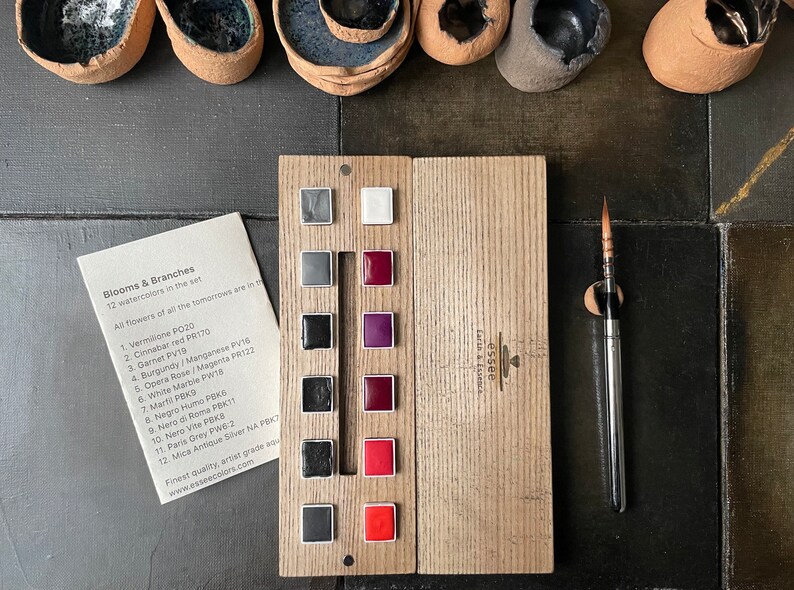 May include: A wooden watercolor palette with 12 color squares, a paintbrush, and a card listing the colors. The palette includes shades of gray, red, and purple. The card reads "Blooms & Branches" and lists the watercolor names.
