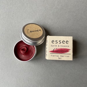 May include: A small, round, silver tin containing a red, waxy substance. The lid of the tin is open and has a brown label with the word "Carmin" written on it. Beside the tin is a small, rectangular, cardboard box with the words "essee Earth & Essence Carmin Tierras Ibericas 5ml" printed on it.
