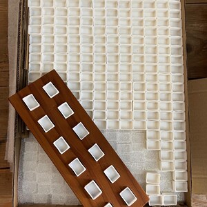 May include: A wooden palette with white watercolor paint wells, alongside a large tray filled with similar empty wells. The palette is angled, showcasing the individual paint compartments. The setup is inside a cardboard box.