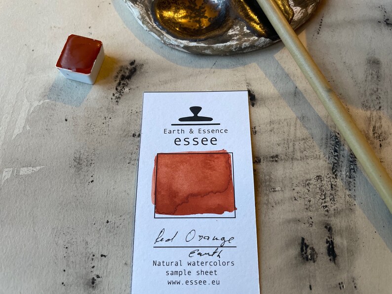 Puede incluir: Una hoja de muestra de pintura de acuarela Tierra naranja roja de la marca Earth & Essence essee. La pintura es de un color naranja-rojo vibrante. La hoja incluye el nombre de la marca, el sitio web y el texto "Muestra de acuarelas naturales".