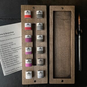 May include: A wooden watercolor set with 12 colorful watercolor paints, a paintbrush, and a card with the text "Blooms & Branches". The set includes various shades of red, pink, gray, and white watercolors.