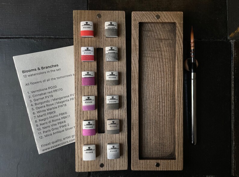 May include: A wooden watercolor set with 12 colorful watercolor paints, a paintbrush, and a card with the text "Blooms & Branches". The set includes various shades of red, pink, gray, and white watercolors.