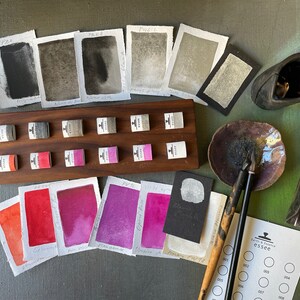 May include: An assortment of watercolor paints and supplies, including sample swatches, paint pans, and calligraphy pens. The color palette ranges from deep blacks and grays to vibrant reds and purples. A wooden holder displays small paint cubes. The image includes the text "essee".