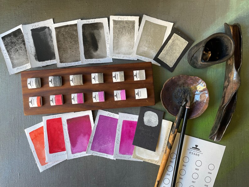 May include: An assortment of watercolor paints and supplies, including sample swatches, paint pans, and calligraphy pens. The color palette ranges from deep blacks and grays to vibrant reds and purples. A wooden holder displays small paint cubes. The image includes the text "essee".