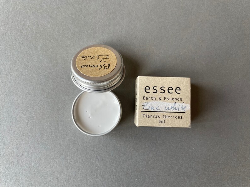 Pu&ograve; includere: Un piccolo contenitore rotondo d'argento con un'etichetta bianca che dice "Blance Zinc" e un'etichetta marrone che dice "essee Earth & Essence Zinc White Tierras Ibericas 5ml".