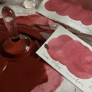 May include: A clear glass bottle stopper with a rounded base sits in a pool of deep red paint. Two watercolour swatches show shades of red and brown, labelled with handwritten text. A palette knife is also visible.