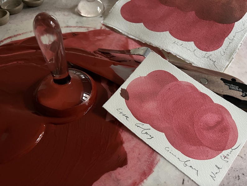 May include: A clear glass bottle stopper with a rounded base sits in a pool of deep red paint. Two watercolour swatches show shades of red and brown, labelled with handwritten text. A palette knife is also visible.
