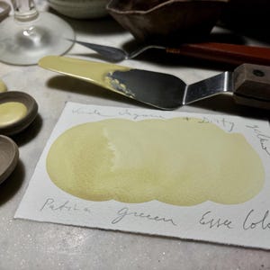 May include: A still life arrangement of art materials. A palette knife with a wooden handle lies near a watercolour swatch. The swatch is a pale yellow colour, with handwritten text reading "Patina Green Essex Colour". Small ceramic bowls are also visible.