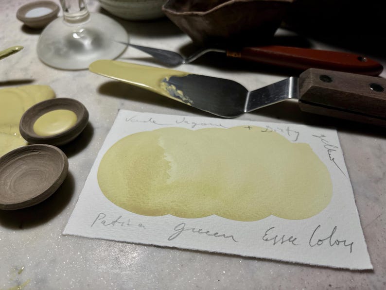 May include: A still life arrangement of art materials. A palette knife with a wooden handle lies near a watercolour swatch. The swatch is a pale yellow colour, with handwritten text reading "Patina Green Essex Colour". Small ceramic bowls are also visible.