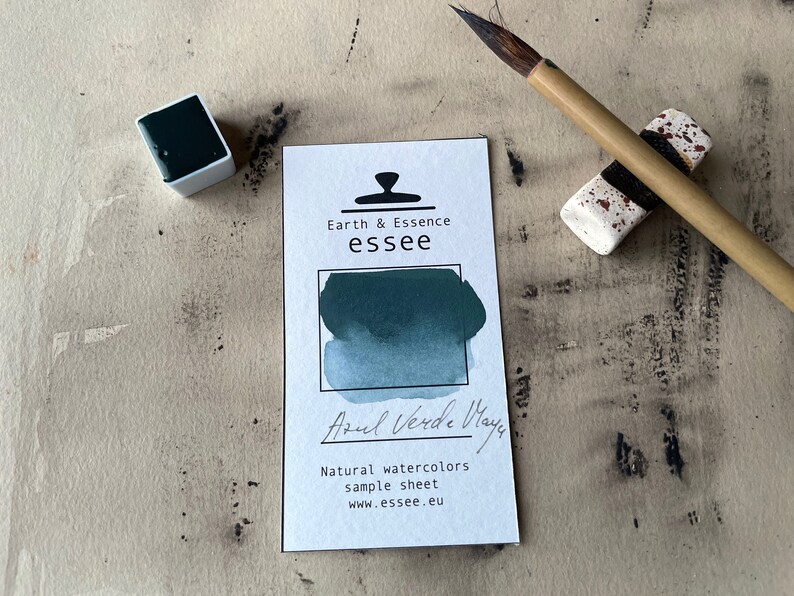 Op de afbeelding: Een aquarelverf-staalkaart met de tekst "Earth & Essence essee" en "Azul Verde Maya". De kaart toont een staal van de aquarelverf in een blauwgroene kleur.