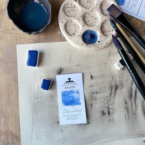 Op de afbeelding: Een close-up van een aquarelverfset met een blauwe aquarelpan, een klein wit kaartje met de tekst "Earth & Essence essee Mage Indige Natural watercolors sample sheet www.essee.eu", en verschillende kwasten op een houten oppervlak.