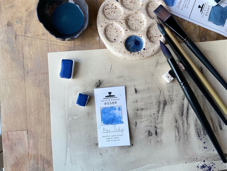 Op de afbeelding: Een close-up van een aquarelverfset met een blauwe aquarelpan, een klein wit kaartje met de tekst "Earth & Essence essee Mage Indige Natural watercolors sample sheet www.essee.eu", en verschillende kwasten op een houten oppervlak.