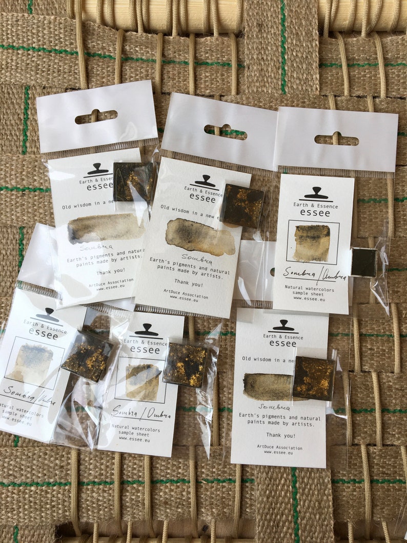 May include: Six small sample watercolour paint squares in clear plastic packaging. Each square is labelled "Sombra" and features a different shade of brown with gold flecks. The packaging includes the brand name "Earth & Essence essee" and the text "Old wisdom in a new way".