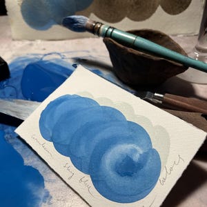 May include: A watercolour painting demonstration featuring a sky blue colour palette. The artwork showcases layered brushstrokes of blue hues, with the words "Sky blue" and "Esser colours" handwritten on the paper. A paintbrush and palette are also visible.