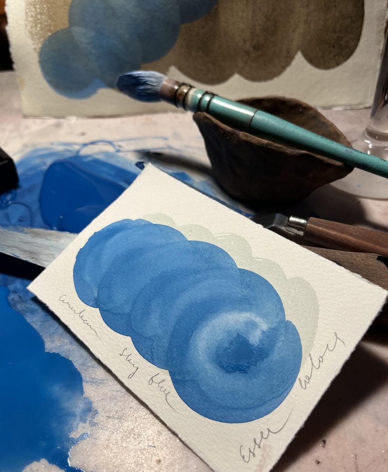 May include: A watercolour painting demonstration featuring a sky blue colour palette. The artwork showcases layered brushstrokes of blue hues, with the words "Sky blue" and "Esser colours" handwritten on the paper. A paintbrush and palette are also visible.