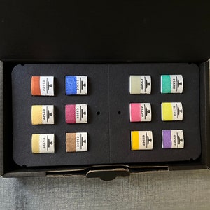 Puede incluir: Una caja negra con un inserto de espuma que contiene 12 peque&ntilde;as bandejas de pintura. Las bandejas est&aacute;n dispuestas en dos filas de seis. Las bandejas est&aacute;n etiquetadas con "essee" y tienen un peque&ntilde;o icono de un pincel. Las bandejas est&aacute;n llenas de diferentes colores de pintura, incluyendo naranja, azul, verde, amarillo, rosa y morado.