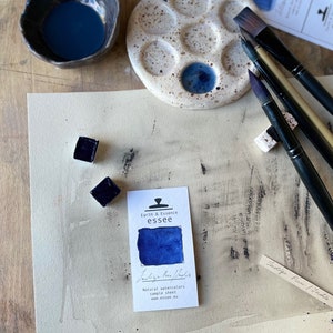 Op de afbeelding: Een aquarelverfset met een blauw pigment, een keramisch palet en kwasten. De verf is gelabeld met "Indigo Raw Powder" en "Earth & Essence essee".