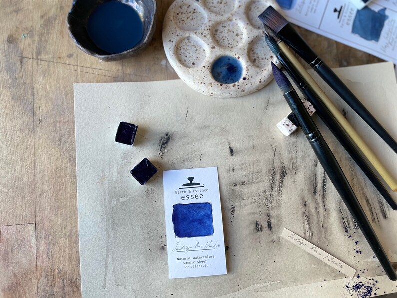 Op de afbeelding: Een aquarelverfset met een blauw pigment, een keramisch palet en kwasten. De verf is gelabeld met "Indigo Raw Powder" en "Earth & Essence essee".