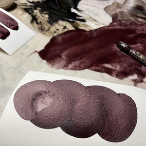 May include: Close-up of watercolour paint swatches in shades of burgundy and plum on white paper. A paintbrush rests on a dark red paint spill. The image showcases the texture and colour variations of the paint.