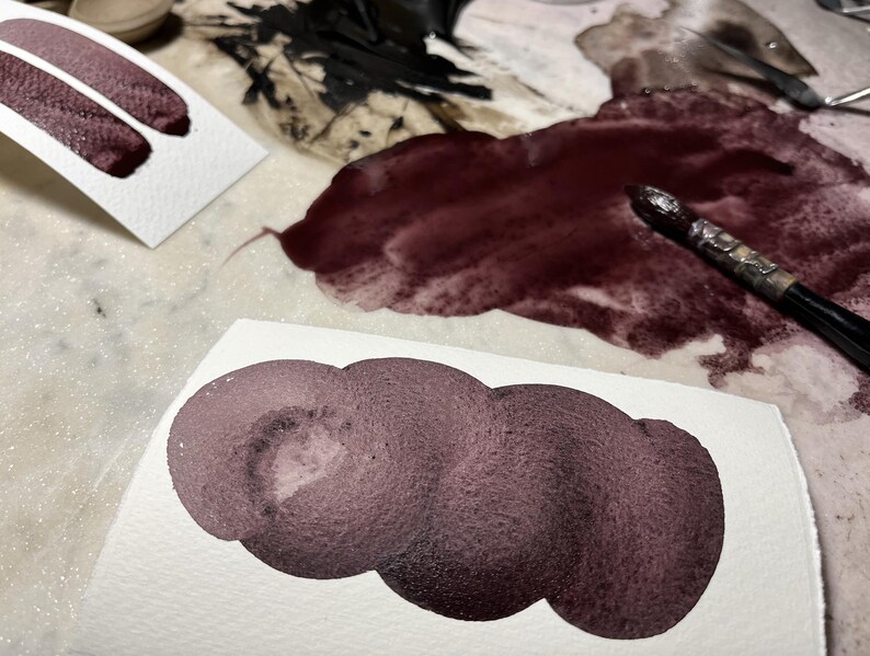 May include: Close-up of watercolour paint swatches in shades of burgundy and plum on white paper. A paintbrush rests on a dark red paint spill. The image showcases the texture and colour variations of the paint.