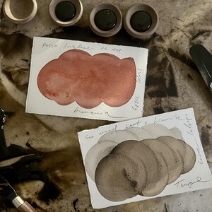 May include: An overhead view of art materials, featuring watercolour paint swatches, paint pots, and a palette knife. The swatches show red and brown hues, with handwritten text. The paint pots hold dark green paint.
