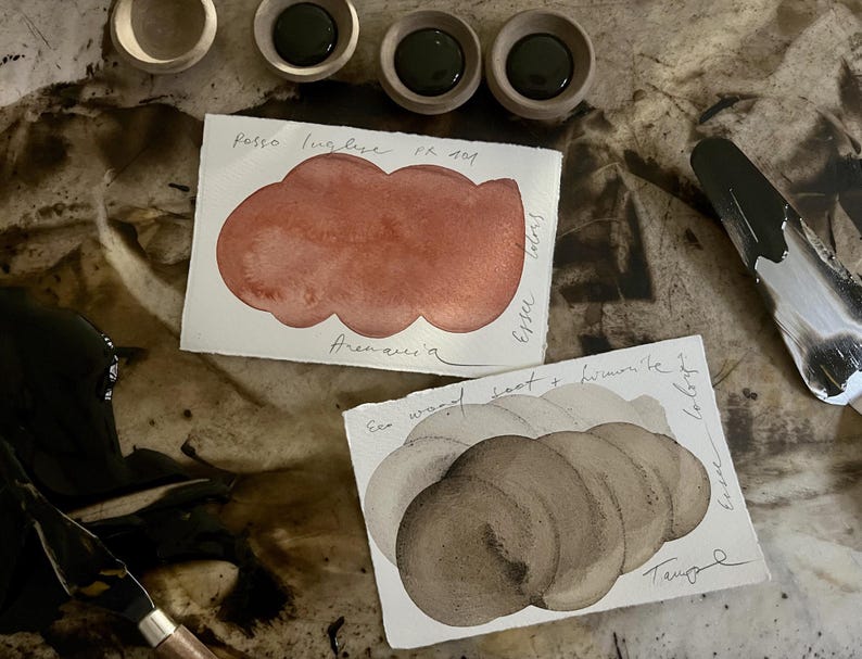 May include: An overhead view of art materials, featuring watercolour paint swatches, paint pots, and a palette knife. The swatches show red and brown hues, with handwritten text. The paint pots hold dark green paint.