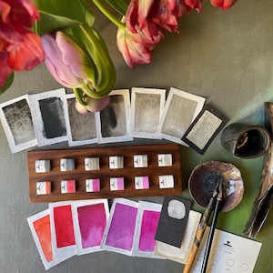 May include: An assortment of art supplies, including ink samples, watercolor swatches, and ink bottles, arranged on a green surface. A wooden tray holds small ink bottles. Watercolor paper samples display various shades of red, pink, and purple. A bouquet of pink and red tulips is in the background.