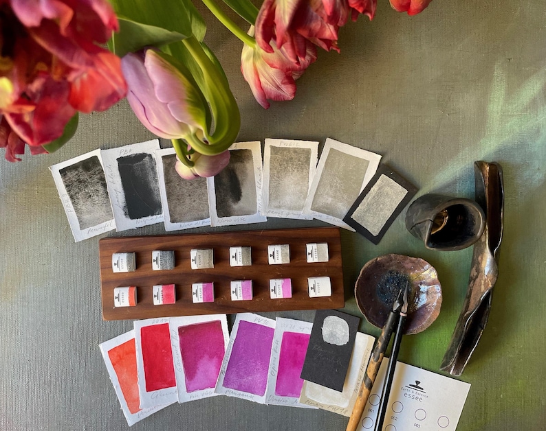 May include: An assortment of art supplies, including ink samples, watercolor swatches, and ink bottles, arranged on a green surface. A wooden tray holds small ink bottles. Watercolor paper samples display various shades of red, pink, and purple. A bouquet of pink and red tulips is in the background.