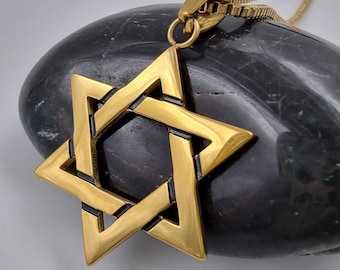 May include: A gold-toned pendant necklace featuring a detailed Star of David design. The pendant is on a black stone surface.
