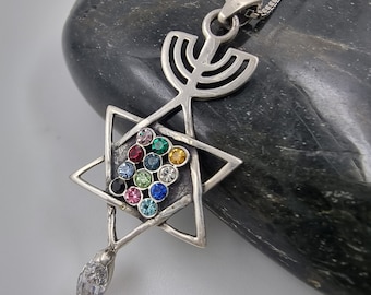 May include: A silver pendant featuring a Star of David with colorful gemstones and a menorah design. The pendant is hanging from a silver chain.