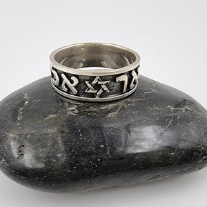 May include: A silver ring featuring a Star of David and Hebrew lettering. The ring is resting on a grey stone.