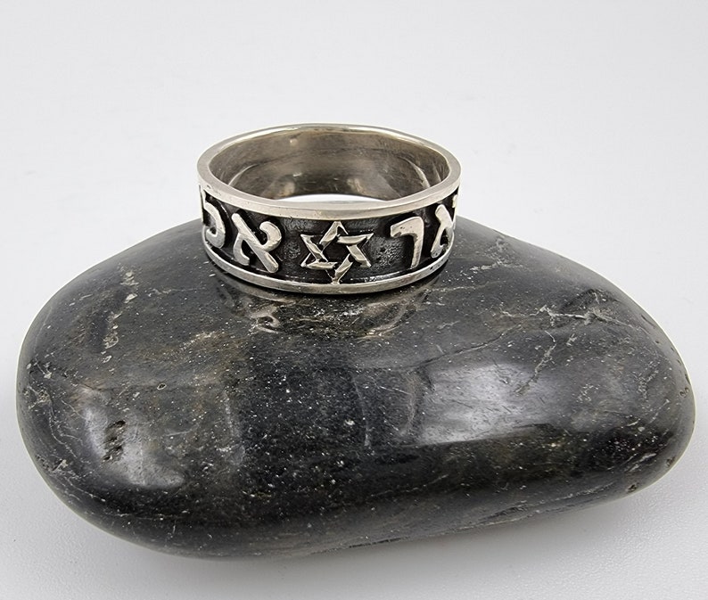 May include: A silver ring featuring a Star of David and Hebrew lettering. The ring is resting on a grey stone.