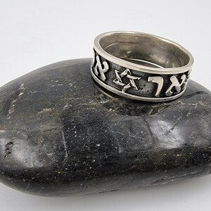 May include: A silver ring featuring Hebrew lettering and a Star of David. The ring is resting on a grey stone.