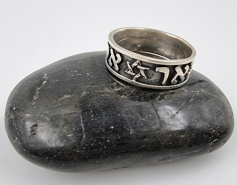 May include: A silver ring featuring Hebrew lettering and a Star of David. The ring is resting on a grey stone.