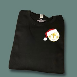 Peut inclure: Sweat-shirt noir avec un motif de Noël blanc et or représentant un bonhomme de neige portant un bonnet de Père Noël. Le texte "The Hitchhiking Ghosts" est imprimé sur le col.