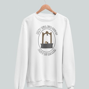 Op de afbeelding: Witte sweatshirt met een afbeelding van een put met een persoon erin en de tekst "DON'T TELL HIM CARLOS DON'T BE CHICKEN" in een zwarte ovaal. De sweatshirt hangt aan een houten hanger.