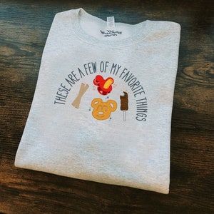 Pode incluir: Camisola cinzenta com o texto "These are a few of my favorite things" num círculo em torno de uma cabeça de Mickey Mouse, um pretzel de Mickey Mouse, um corn dog de Mickey Mouse e macarrão em forma de Mickey Mouse.