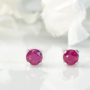 May include: A pair of round, faceted, deep pinkish-red ruby stud earrings set in four-prong silver metal settings. The stones exhibit a clear, polished surface with visible internal facets that catch the light, creating a bright, reflective appearance.