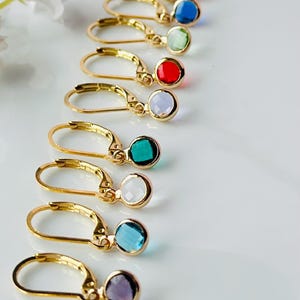 Puede incluir: Una colección de pendientes dorados con cierre de palanca, cada uno con una piedra preciosa de diferente color. Las piedras preciosas incluyen tonos de azul, verde, rojo, morado y blanco. Los pendientes están dispuestos en fila.