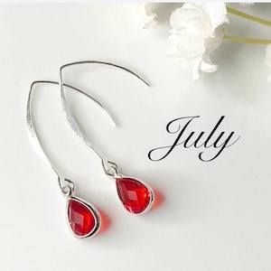 May include: A pair of silver-toned drop earrings featuring faceted, teardrop-shaped red stones. The earrings have curved wire hooks and the word July is written in a black cursive script font next to the jewelry pieces.