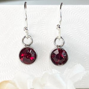 May include: A pair of dangle earrings with a silver-colored metal setting. Each earring features a round, faceted, deep red gemstone. The earrings hang from a simple hook.